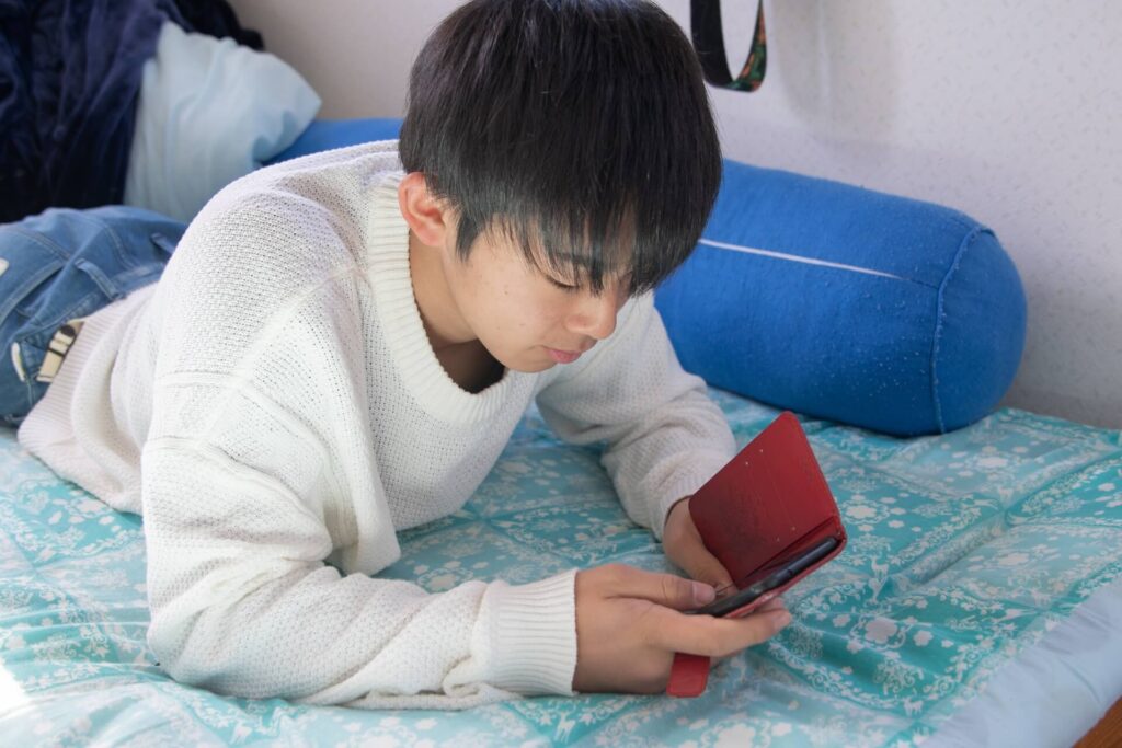 男子高校生が寝そべりなっがらスマートフォンを操作している写真です。